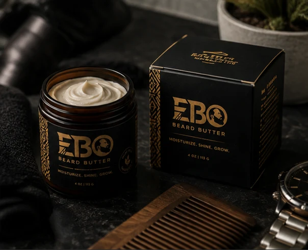 An open EBO beard butter jar beside its black-and-gold box, a wooden comb and a watch on dark stone.